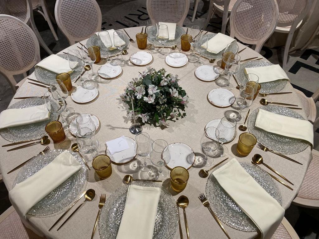 Mesa redonda de boda vestida en blanco con cubertería dorada, cristalería y centro floral