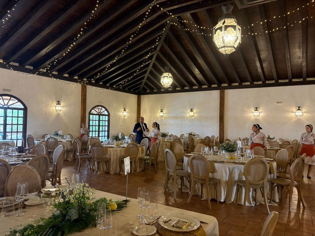Salón de una hacienda de Sevilla montado para un banquete de boda, con mesas redondas, techo de vigas de madera y guirnaldas de luces